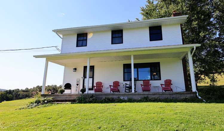 Modern country home with white siding and black windows, featuring a front porch with red chairs, set in a green landscape.